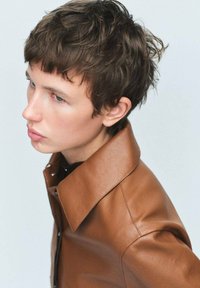 Chaqueta de cuero marrón con un amplio collar, superficie texturizada y una silueta ajustada, usada sobre una prenda negra. Primer plano del perfil del modelo.