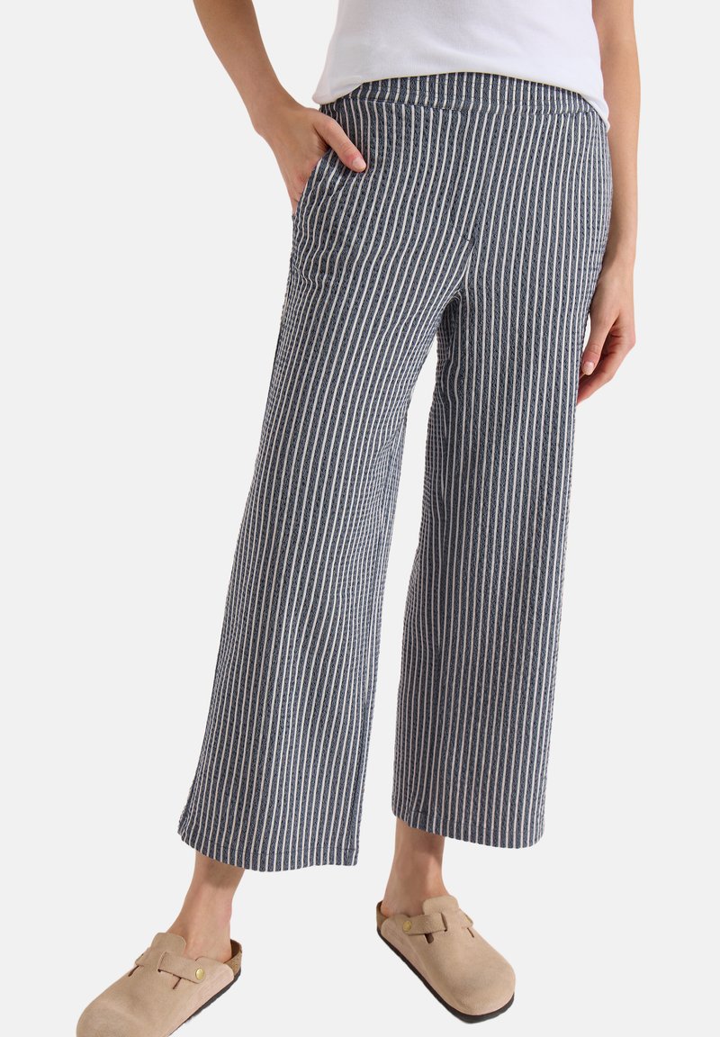 Femme portant un pantalon large à rayures verticales bleu marine et blanc avec poches, associé à des sabots beiges à enfiler et un haut blanc.