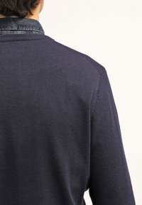 Granatowy sweter o teksturowanym splotem, z kontrastowym kołnierzem z denimu. Widok z tyłu ukazuje subtelne detale szwów na ramionach.