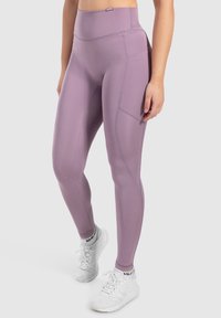 Hoge taille legging in lichtpaars, gemaakt van rekbare stof. Bevat een zijzak en naaddetails. Gecombineerd met witte sportschoenen.