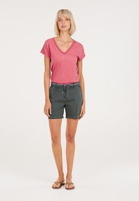 T-shirt rosa a scollatura a V abbinato a pantaloni corti verde scuro; i pantaloni corti presentano una cintura con un bottone e un cordino, e sono arrotolati all'orlo.
