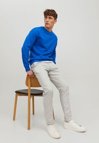 Sweatshirt bleu, pantalon gris clair et baskets blanches. Assis sur une chaise en bois avec un siège noir. Design simple, sans motifs notables.