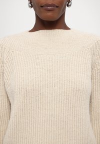 Gros plan d'une personne portant un pull crème en maille côtelée et des boucles d'oreilles argentées en forme d'anneau, avec les lèvres visibles mais les yeux hors cadre.