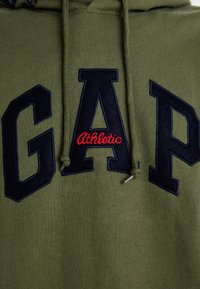 Hoodie vert olive avec un grand logo "GAP" bleu marine et le texte "Athletic" en rouge. Le matériau est doux avec une finition texturée, cordons de serrage inclus.