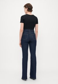 Person with short curly hair wearing black fitted short-sleeve top, high-waisted dark blue jeans, and black heeled shoes standing facing away.