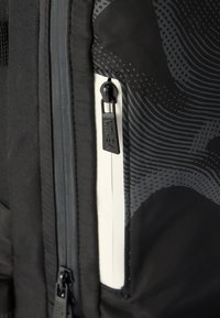 Mochila de tecido preto com padrões abstratos cinzentos, apresentando um bolso com fecho de correr, puxador preto do fecho e superfície texturizada. Detalhe branco visível.