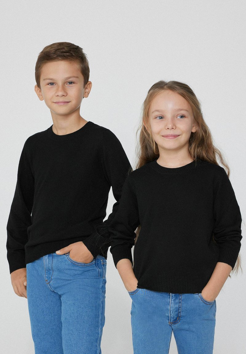 Zwei Kinder stehen nebeneinander, tragen schwarze Strickpullover und blaue Jeans. Die Pullover haben einen runden Ausschnitt und gerippte Bündchen.