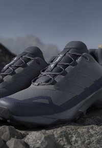 Σκούρα γκρι παπούτσια trail running Adidas Terrex σε πετρώδες έδαφος με ορεινό τοπίο κάτω από καθαρό ουρανό.