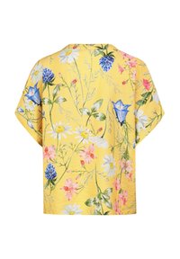 Gelbe Blumenbluse mit kurzen Ärmeln, die weiße Gänseblümchen, rosa und blaue Blumen sowie grüne Blätter auf einem strukturierten Hintergrund zeigt.