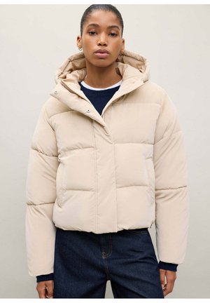 Une femme porte une doudoune courte beige à capuche sur un haut bleu marine et un jean foncé, devant un fond clair uni.