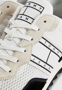 Zapatilla blanca con materiales de malla y ante, detalle de logotipo a rayas en negro y blanco, cordones planos blancos y suela negra texturizada.