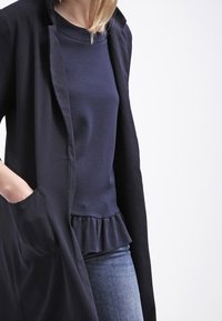 Haut peplum marine sous un long manteau noir à revers, avec des poches et une texture lisse, associé à un jean en denim.