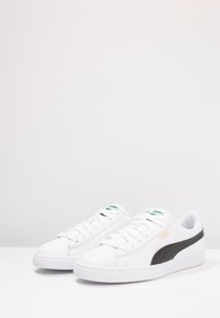 Baskets blanches avec dessus en cuir lisse, bande noire sur le côté et semelle en caoutchouc. Caractéristiques : bout rond et fermeture à lacets avec des accents verts.