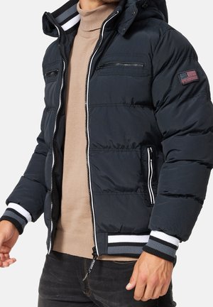 Man draagt een zwart gewatteerd jack met gestreepte manchetten en een patch van de Amerikaanse vlag, over een beige coltrui.