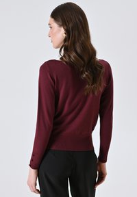 Bordeauxkleurige sweater met een getailleerd ontwerp, lange mouwen en knoopaccenten op de manchetten. Zachte textuur, ronde halslijn en geribbelde zoom.