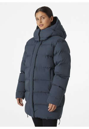 Donkerblauwe pufferjack met een hoge kraag en grote capuchon. Voorzien van een verticale gestikte ontwerp en elastische manchetten, ontworpen voor warmte.