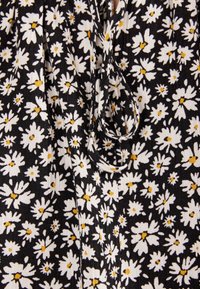 Tessuto nero con margherite bianche e centri gialli. Presenta una texture arricciata e un delicato dettaglio con fiocco sul collo.