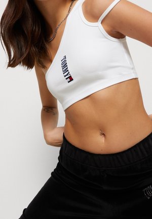 Woman wearing a white Tommy crop top with a necklace and black pants, showing a tattoo on left arm and a belly button ring.