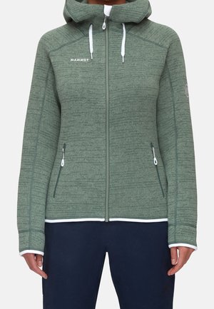 Zielona kurtka z kapturem na zamek błyskawiczny z białymi sznurkami, kieszeniami na zamek po bokach i logo „Mammut” na lewej piersi, noszona z granatowymi spodniami.