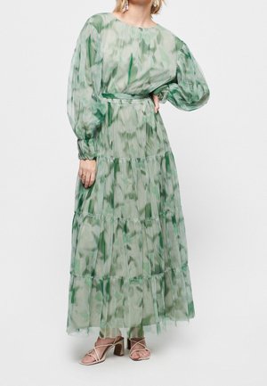 Femme portant une longue robe verte à motif tie-dye, manches longues bouffantes, taille cintrée et jupe à volants, associée à des talons beige à brides.