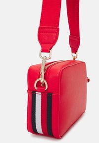 Punainen synteettisestä nahasta valmistettu crossbody-laukku suorakaiteen muotoisena, kultaiset yksityiskohdat ja raidoitettu korostus (punainen, musta, valkoinen) sivussa.