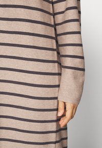 Proužkovaný svetr v béžové barvě s tmavě námořnickými pruhy; měkká textura, dlouhé rukávy, volný střih, viditelný šev na manžetě.