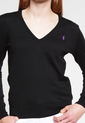 Pull noir à col en V avec logo violet brodé sur le côté gauche de la poitrine, porté par une personne les mains sur les hanches.