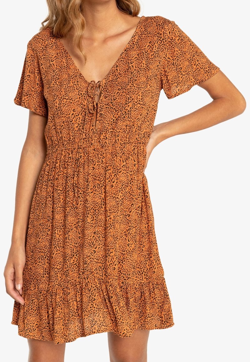 Femme portant une robe orange à manches courtes avec imprimé léopard, col en V et détail noué, debout avec une main sur la hanche.