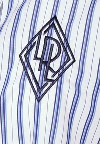 Vit skjorta med vertikala blå ränder och en svart broderad geometrisk logotyp med bokstäver inuti en diamantform.