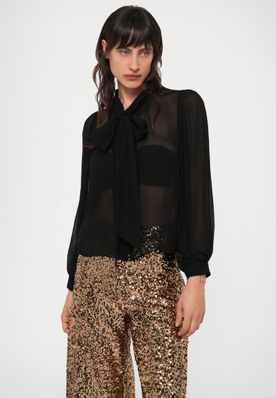 Bluză neagră din material transparent, cu mâneci lungi și detaliu cu fundiță la guler, asortată cu pantaloni aurii cu paiete pentru un contrast textural.
