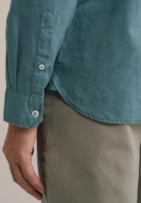 Hosszú ujjú ing tompa teal színű anyagból, gombos mandzsettákkal. Puha tapintás, ívelt alsó szegély és kontrasztos fehér gombok. Bézs nadrággal párosítva.