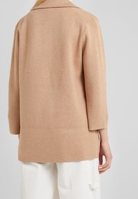 Beige kardigan s dlouhým rukávem a límcem, s texturovaným pleteným designem a bočními rozparky pro snadný pohyb. Zadní pohled ukazuje jednoduchost stylu.
