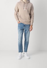 Sweat à capuche beige avec poche avant et logo, associé à un jean bleu clair et des baskets blanches. Tissu décontracté, coupe confortable, design minimaliste.