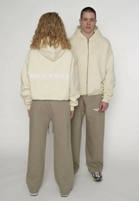 Flødefarvet hættejakke med lynlås og "MULTIPLY"-logo; kombineret med beige, vidde bukser. Blødt stof, afslappet pasform og diskret branding.