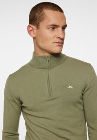 Olivgrön tröja med hög krage och halvmiljö. Har en ribbad textur och en vit logotyp på vänster bröstområde.