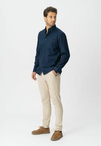 Man die staat met zijn handen in zijn zakken, gekleed in een marineblauwe knoophemd, beige broek en bruine suède schoenen, tegen een eenvoudige witte achtergrond.