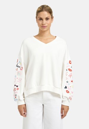 Woman wearing a white V-neck sweatshirt with colorful embroidered symbols and words on both sleeves, paired with white pants.