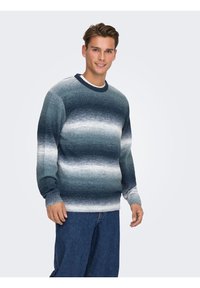 Jeune homme souriant, portant un pull rayé à dégradé bleu et gris et un jean bleu, debout devant un fond uni clair.