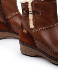 Botas de cuero marrón hasta el tobillo con un ribete de borrego, costura en zigzag, cremallera lateral y suela de corcho en cuña. Detalle de marca visible en el lateral.