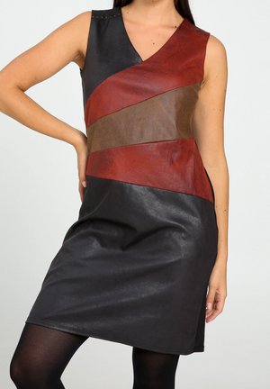 Mouwloze jurk met diagonale strepen in zwart, donkerrood en bruin kunstleer, gedragen met zwarte panty's door een persoon die met één hand op de heup staat.