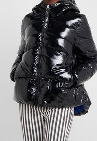 Osoba w błyszczącej czarnej kurtce puchowej z kapturem i metką suwaka "PINKO", w zestawieniu z czarno-białymi spodniami w pionowe paski.