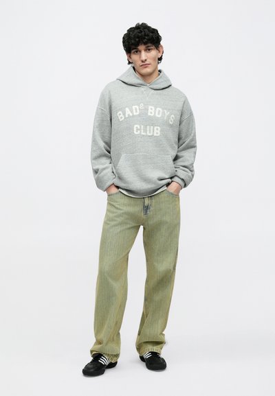 Giovane con capelli ricci che indossa una felpa grigia con la scritta "Bad Boys Club", jeans larghi verde chiaro e sneakers nere con strisce bianche, in piedi in atteggiamento rilassato.