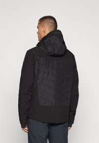 Schwarze Jacke mit einem gepolsterten Rückenteil mit Chevron-Steppung, einer Kapuze und einem glatten Außenmaterial. Mit Reißverschlusstaschen.