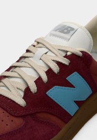 Baskets rouges et bordeaux avec lacets beiges, logo latéral bleu, étiquette blanche sur la languette et semelle marron, vues en gros plan sous un angle.