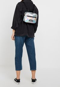 Mochila Adidas holográfica con un logo negro, que cuenta con correas ajustables, un bolsillo frontal con cremallera y forma rectangular. Lleva puesta en la espalda.