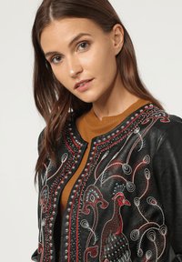 Mujer con cabello largo y castaño vistiendo una chaqueta de cuero negra con un pavo real bordado en rojo y blanco, y tachuelas decorativas sobre una blusa marrón.