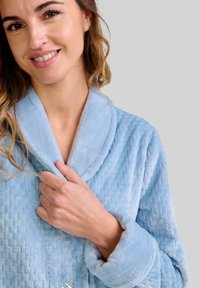 Mujer sonriendo, con un albornoz de textura azul claro, sosteniendo el cuello cerca de su cuello con la mano derecha.