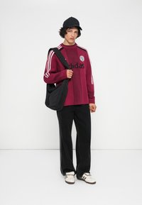 Jauns cilvēks tērpies maroon krāsas Adidas džemperī, melnās biksēs, baltos sporta apavos un melnā cepurē, stāv ar melnu plecu somu pretī baltai fona.