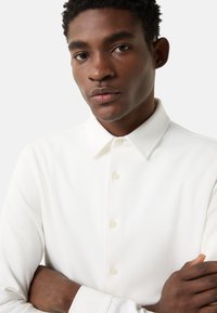 Weißes Langarmhemd mit Knopfkragen, das eine glatte Textur und einen dezenten Glanz aufweist. Es hat passende Knöpfe und eine körpernahe Passform.