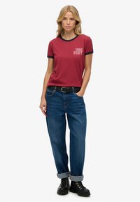 Camiseta roja con cuello en contraste azul oscuro, que presenta un gráfico de "1982 SDRY". Combinada con jeans azules, con dobladillos en los tobillos, y botas negras.